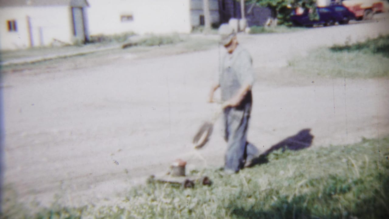 The man pauses or shifts position beside a vintage push mower on a grass strip in the Saint Francis residential neighborhood.