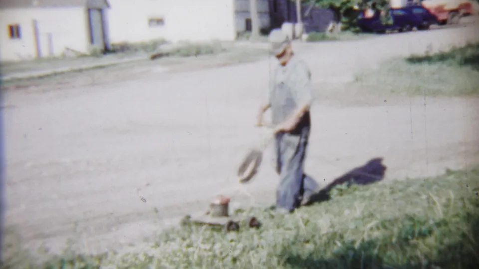 The man pauses or shifts position beside a vintage push mower on a grass strip in the Saint Francis residential neighborhood.