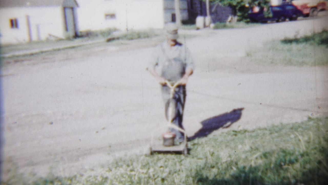 An older man in work clothes faces the home-movie camera while holding the mower handle beside a sunlit residential street.