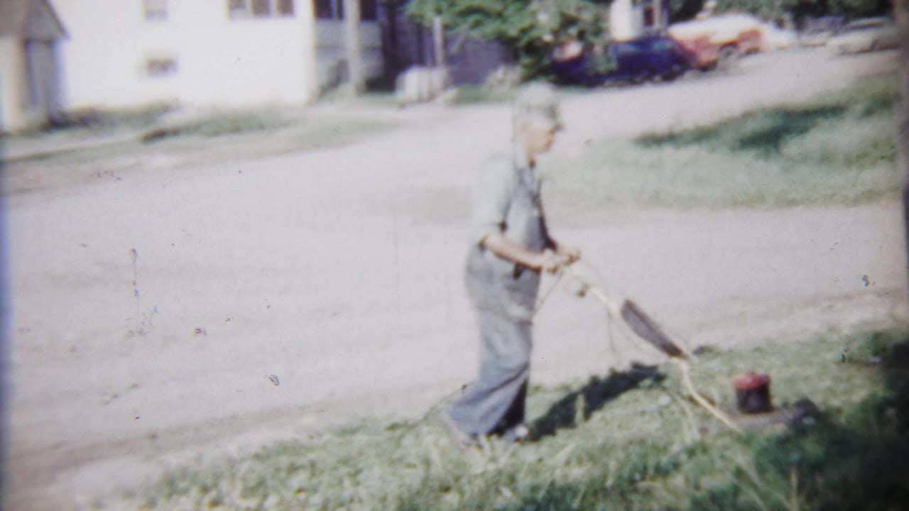 The man walks across the grass near the mower, with Saint Francis houses and parked cars visible beyond the residential street.
