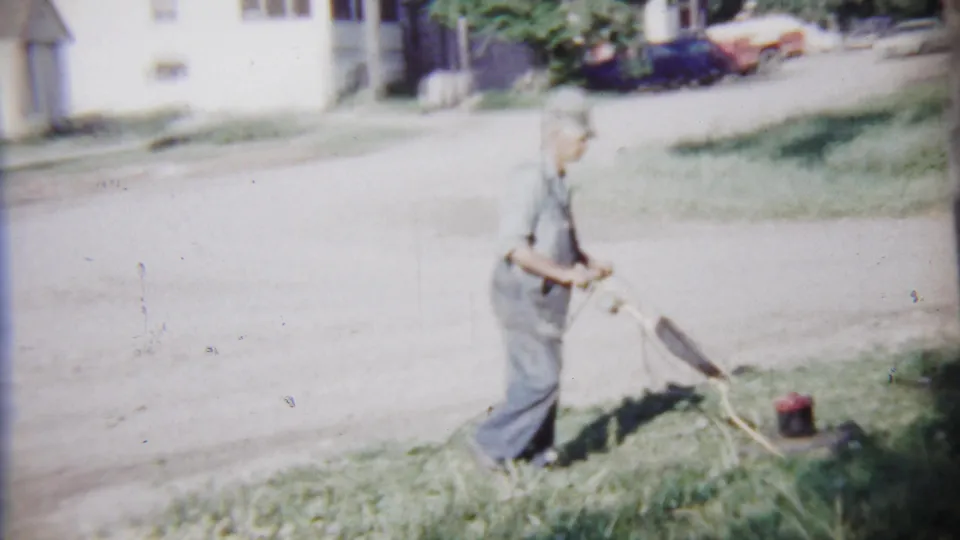 The man walks across the grass near the mower, with Saint Francis houses and parked cars visible beyond the residential street.
