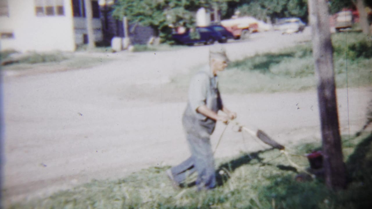 Man in overalls sweeping beside a roadside near a utility pole and parked cars.