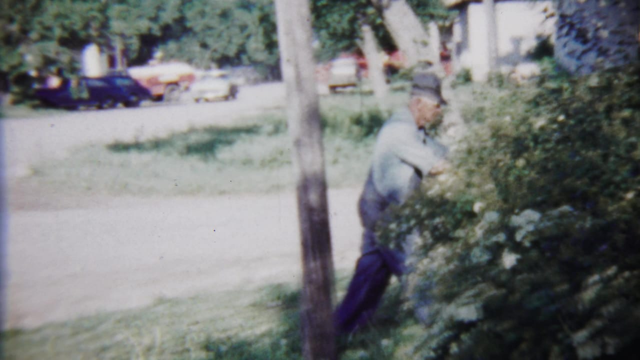 The home-movie frame shows a man working near flowering shrubs along a roadside in Saint Francis, Kansas, with vintage cars beyond the yard.