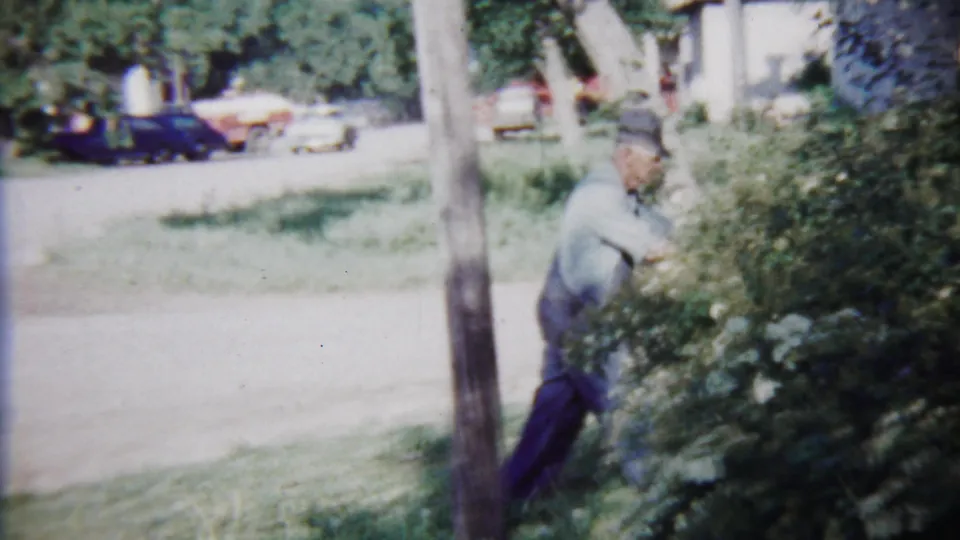 The home-movie frame shows a man working near flowering shrubs along a roadside in Saint Francis, Kansas, with vintage cars beyond the yard.