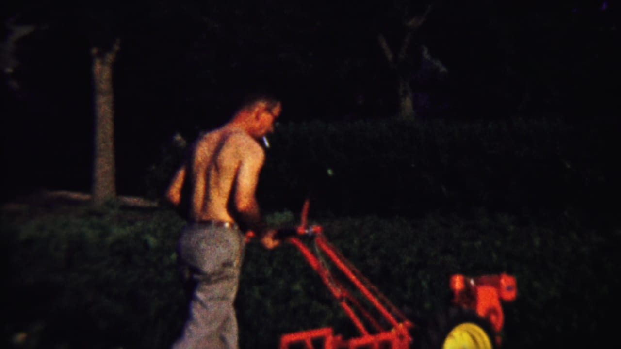 At night, a shirtless man operates a red power tiller in a dark yard, continuing the domestic work sequence from the 1958 home movie.