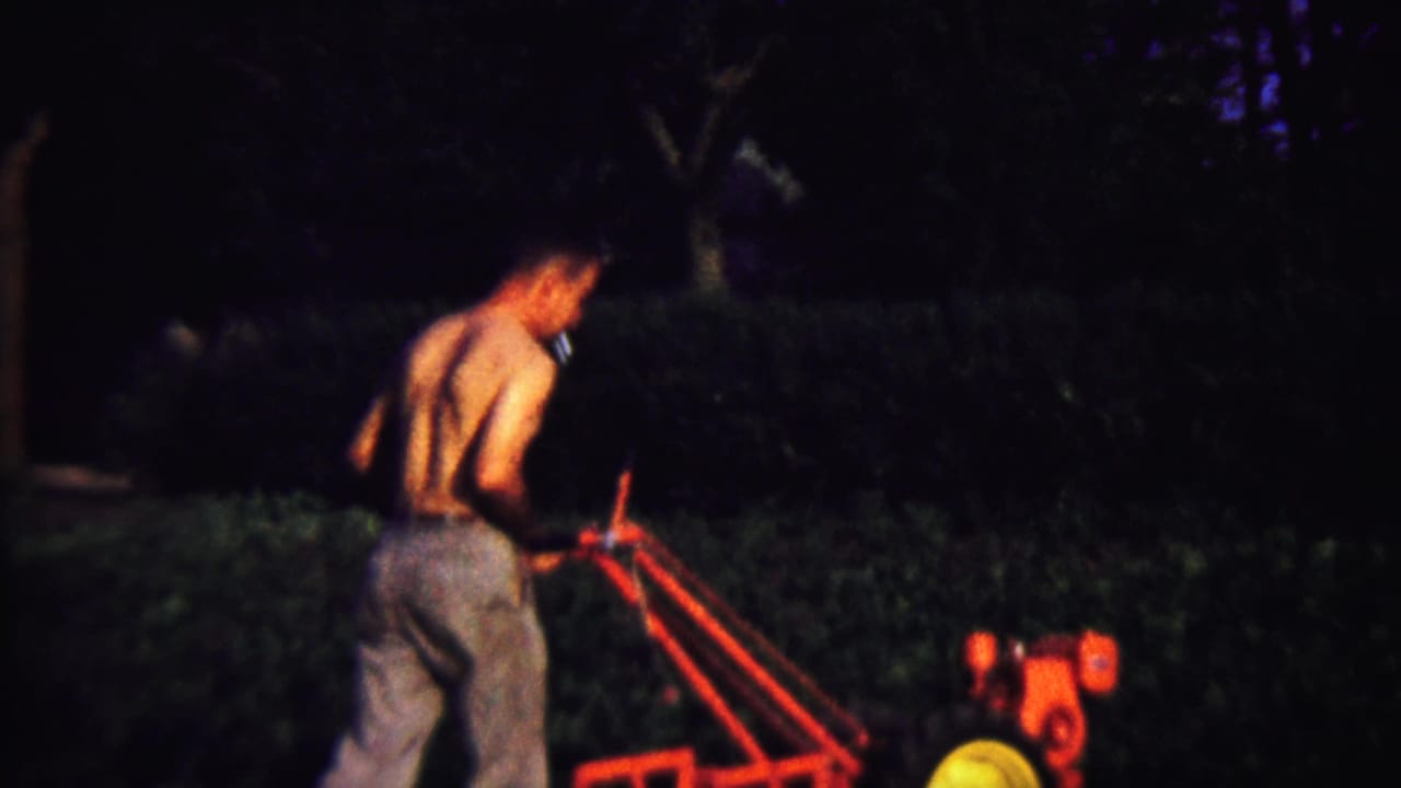 Shirtless man in jeans operates a red hand tiller in a dark yard.
