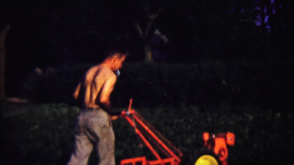 In likely Saint Francis, Kansas, 1958 footage, a shirtless man guides a red hand tiller through a dim backyard.
