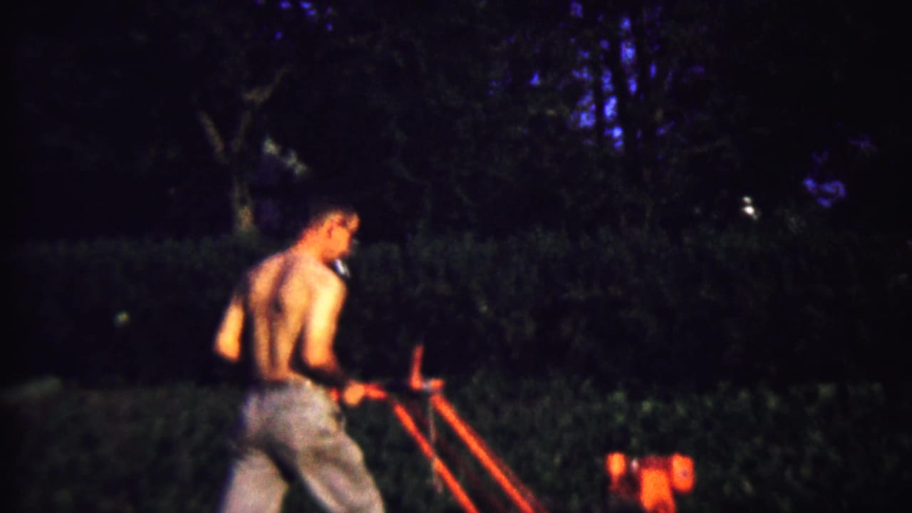 A shirtless man in jeans walks behind a red garden tiller in dim 1958 Kansas home-movie footage.