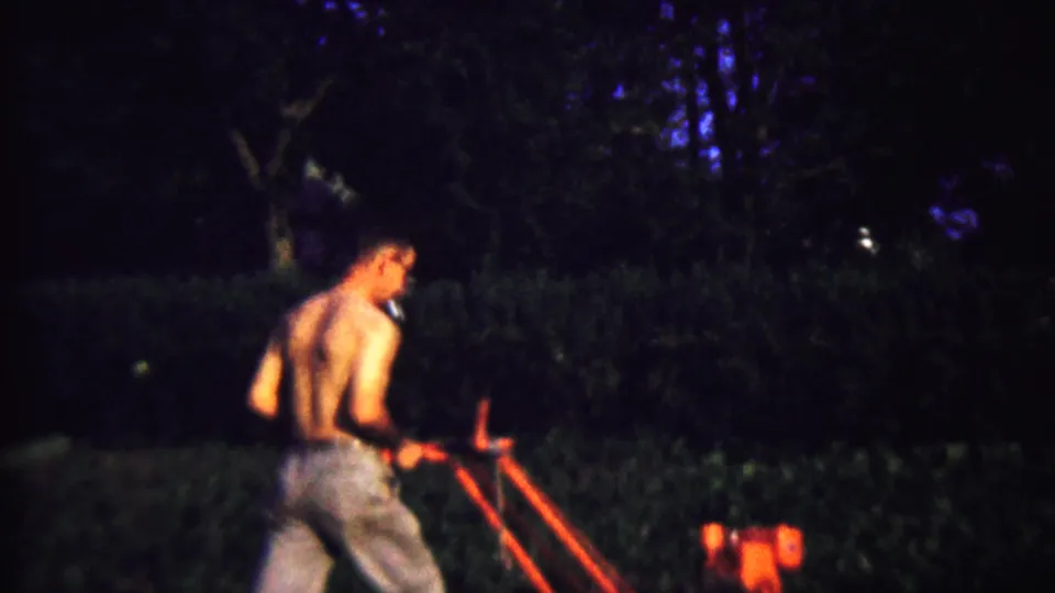 A shirtless man in jeans walks behind a red garden tiller in dim 1958 Kansas home-movie footage.