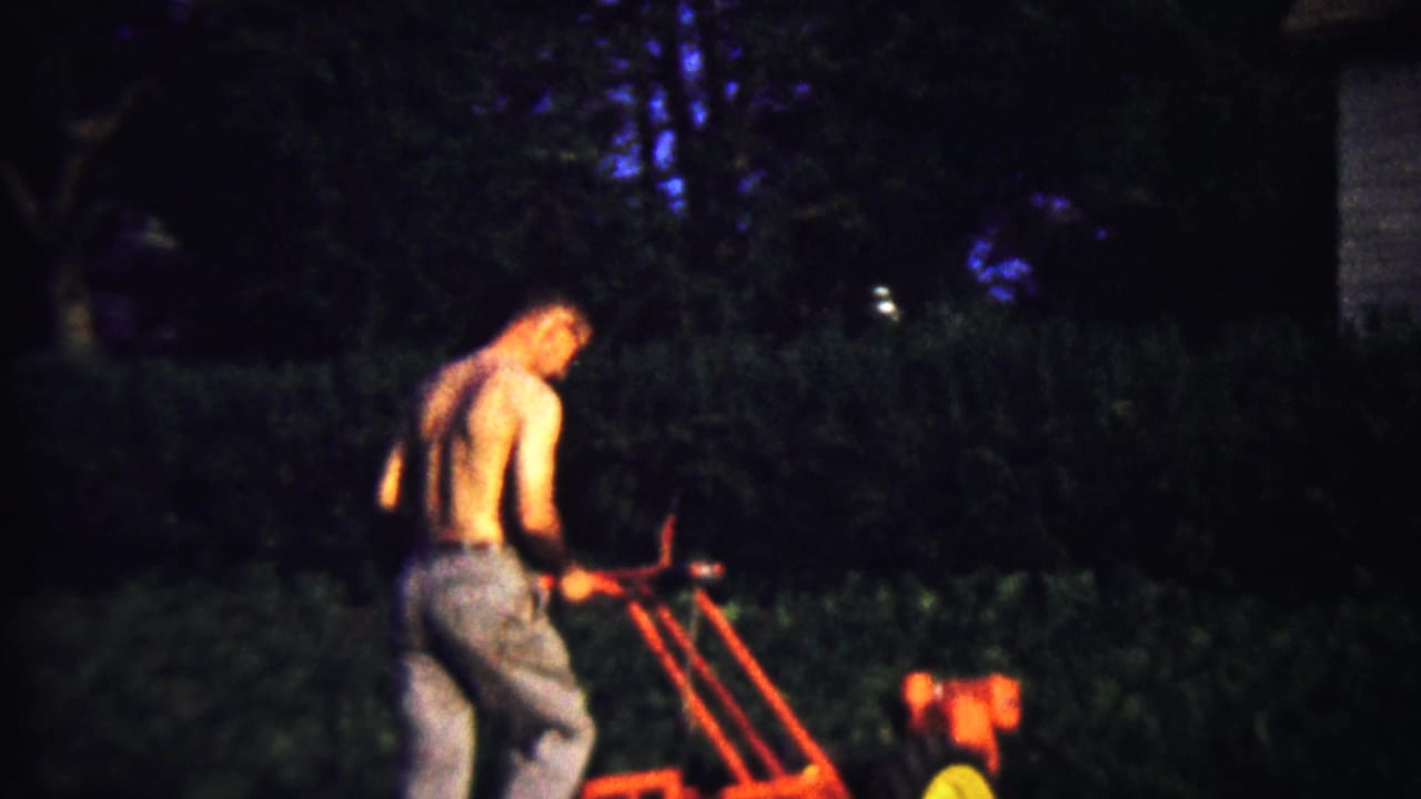 The home-movie frame shows the man leaning into the red tiller, with trees and hedges behind him.