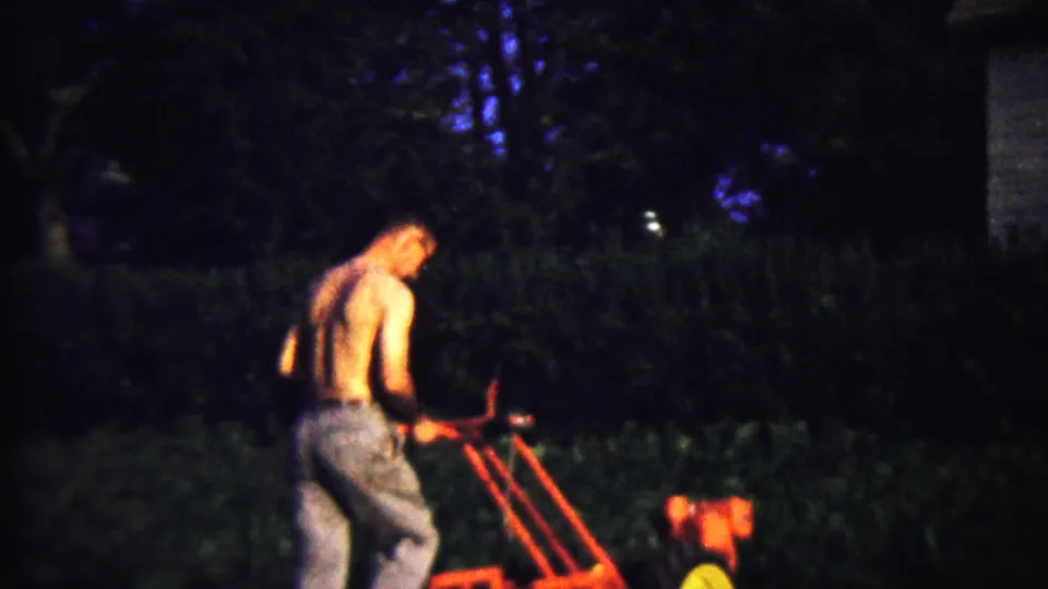 The home-movie frame shows the man leaning into the red tiller, with trees and hedges behind him.
