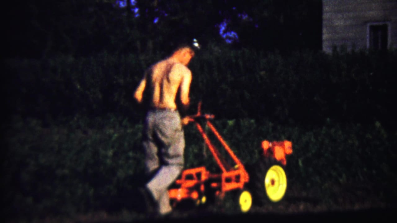 A shirtless man guides the red tiller near dark hedges, with part of a house visible at the right edge.