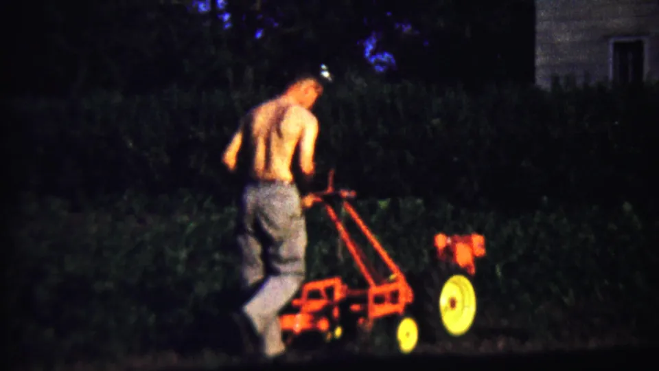 A shirtless man guides the red tiller near dark hedges, with part of a house visible at the right edge.