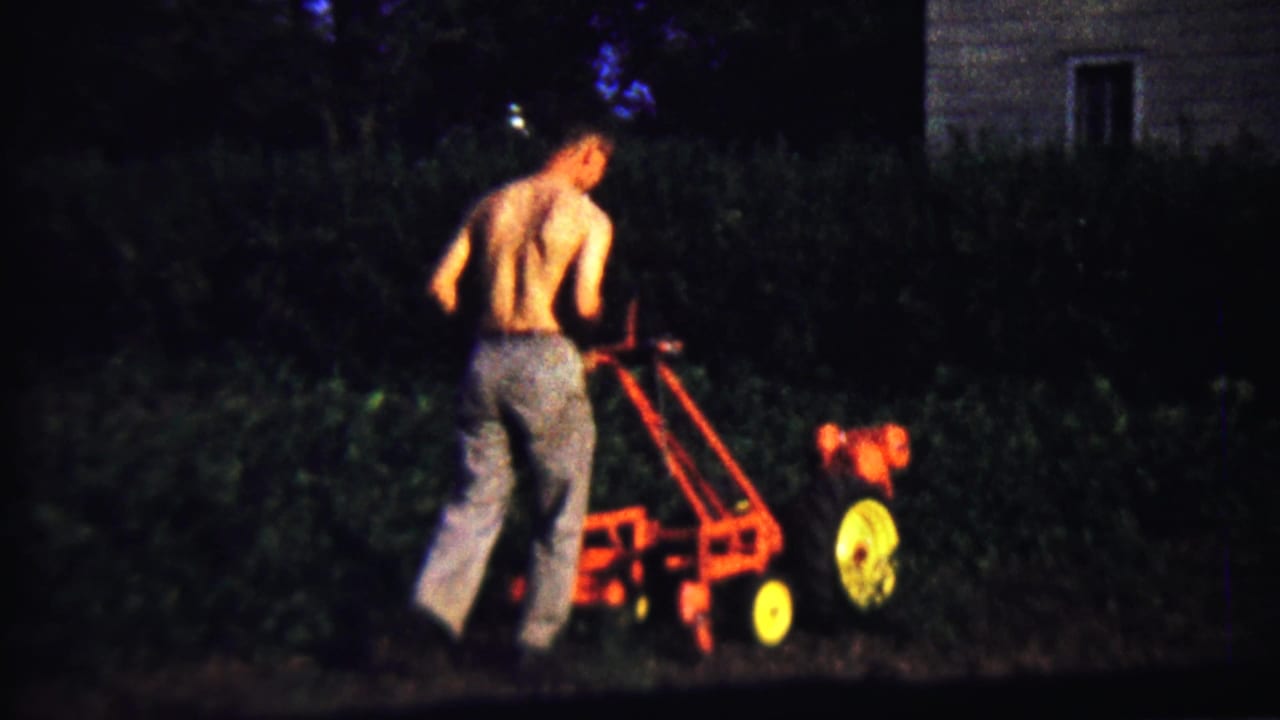 Shirtless man pushing a red and yellow garden tiller beside a dark hedge and house.