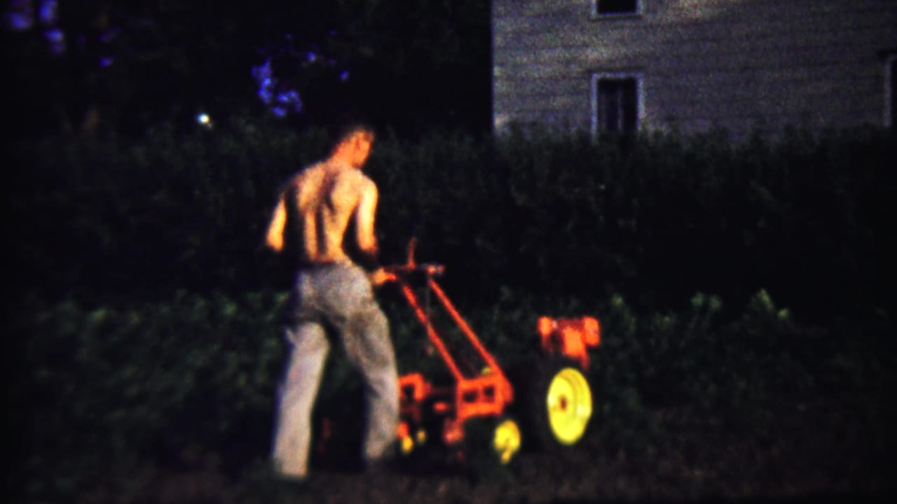 The man continues guiding the walk-behind tiller along the backyard edge, with dark plants and a house exterior behind him.