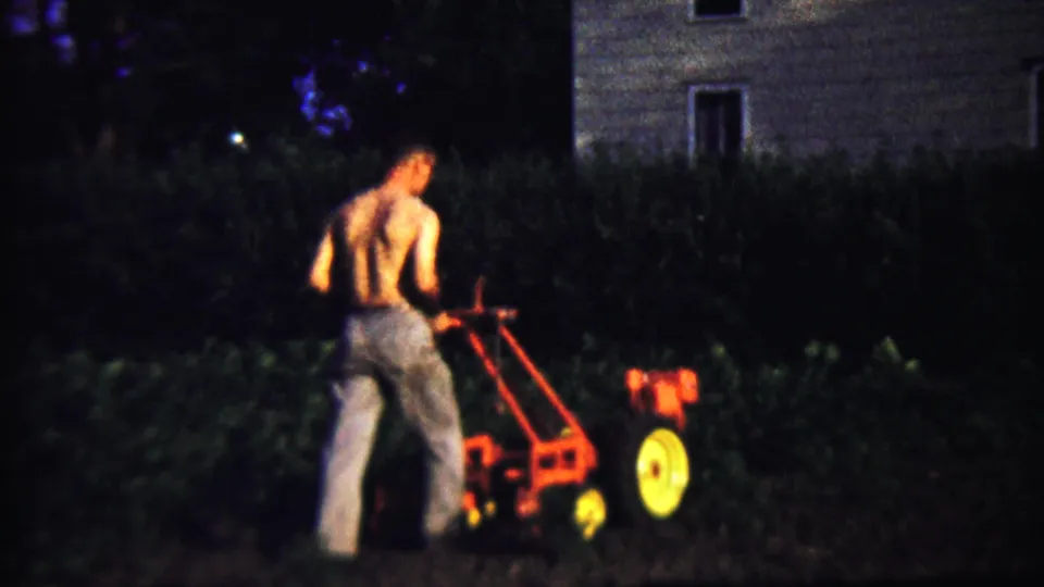 The man continues guiding the walk-behind tiller along the backyard edge, with dark plants and a house exterior behind him.