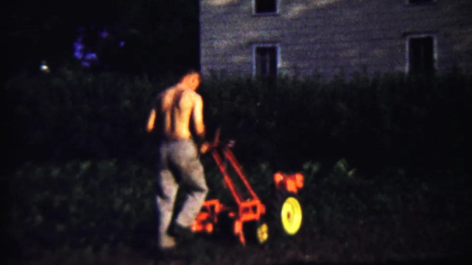 A soft 8mm frame shows the man working the red tiller near dark vegetation and a house in Saint Francis, Kansas.