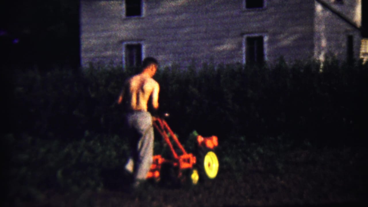 Shirtless man pushes a small red tiller in a dark garden near a house.