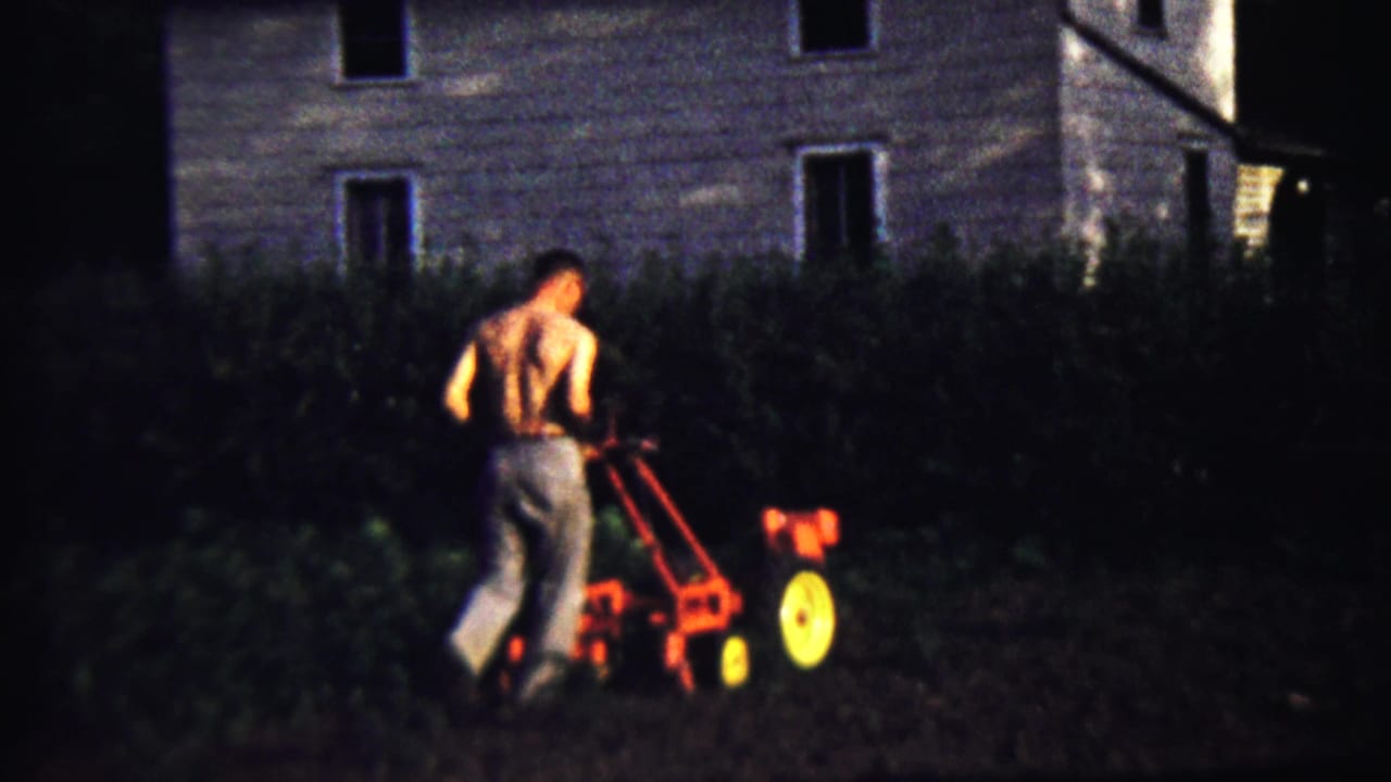 The 1958 Kansas frame shows the man from behind as he operates a red walk-behind tiller near a hedge and house exterior.