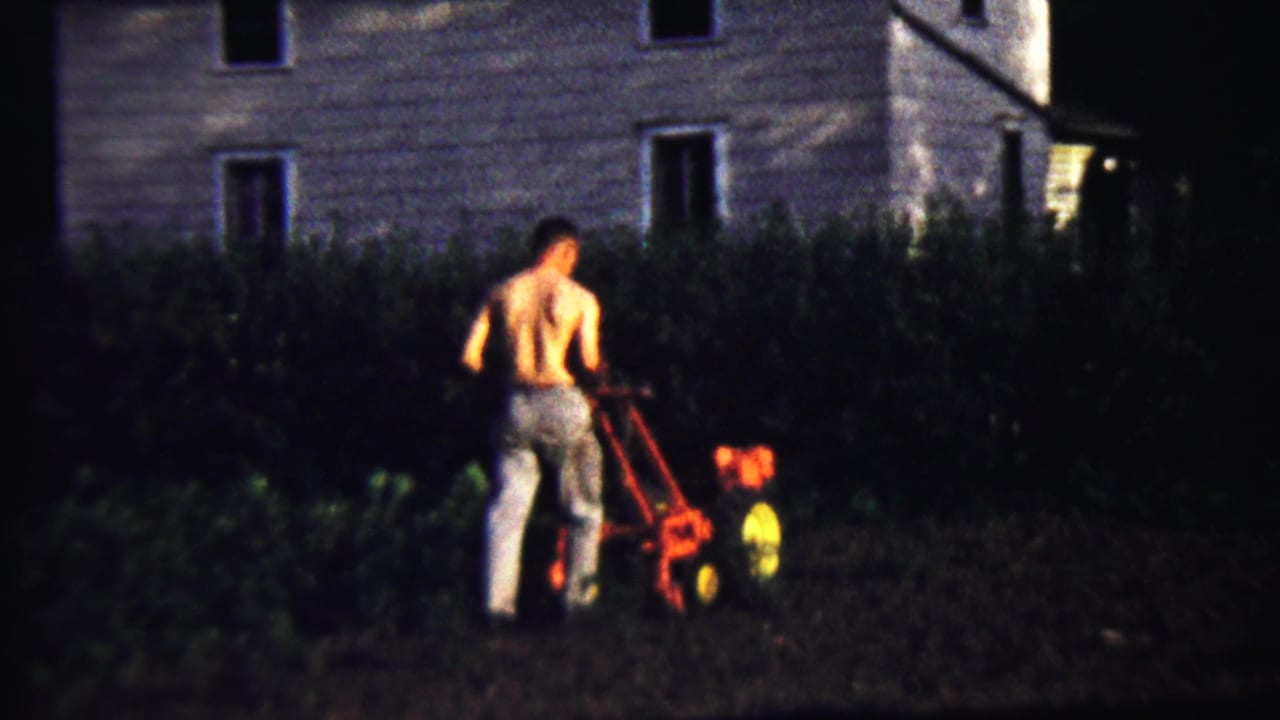 A shirtless man in trousers continues tilling a garden at night in Saint Francis, Kansas, with a house wall behind the hedge.