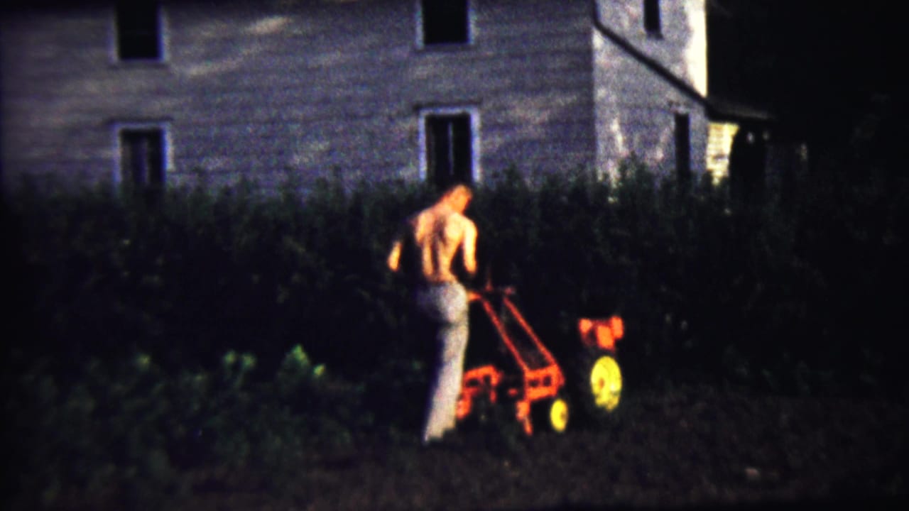 Shirtless man pushes a red garden tiller near a house at night.