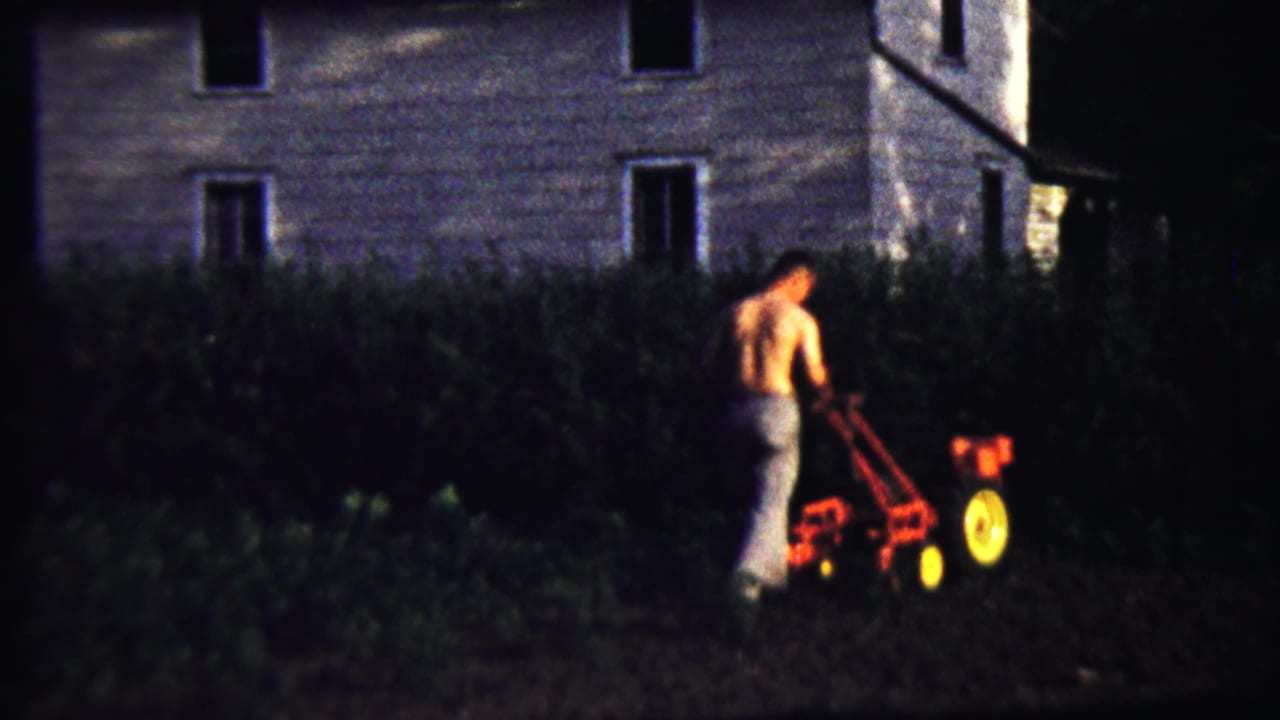 The man continues using a small red tiller in a low-lit garden area in Saint Francis, Kansas, with the house wall behind him.