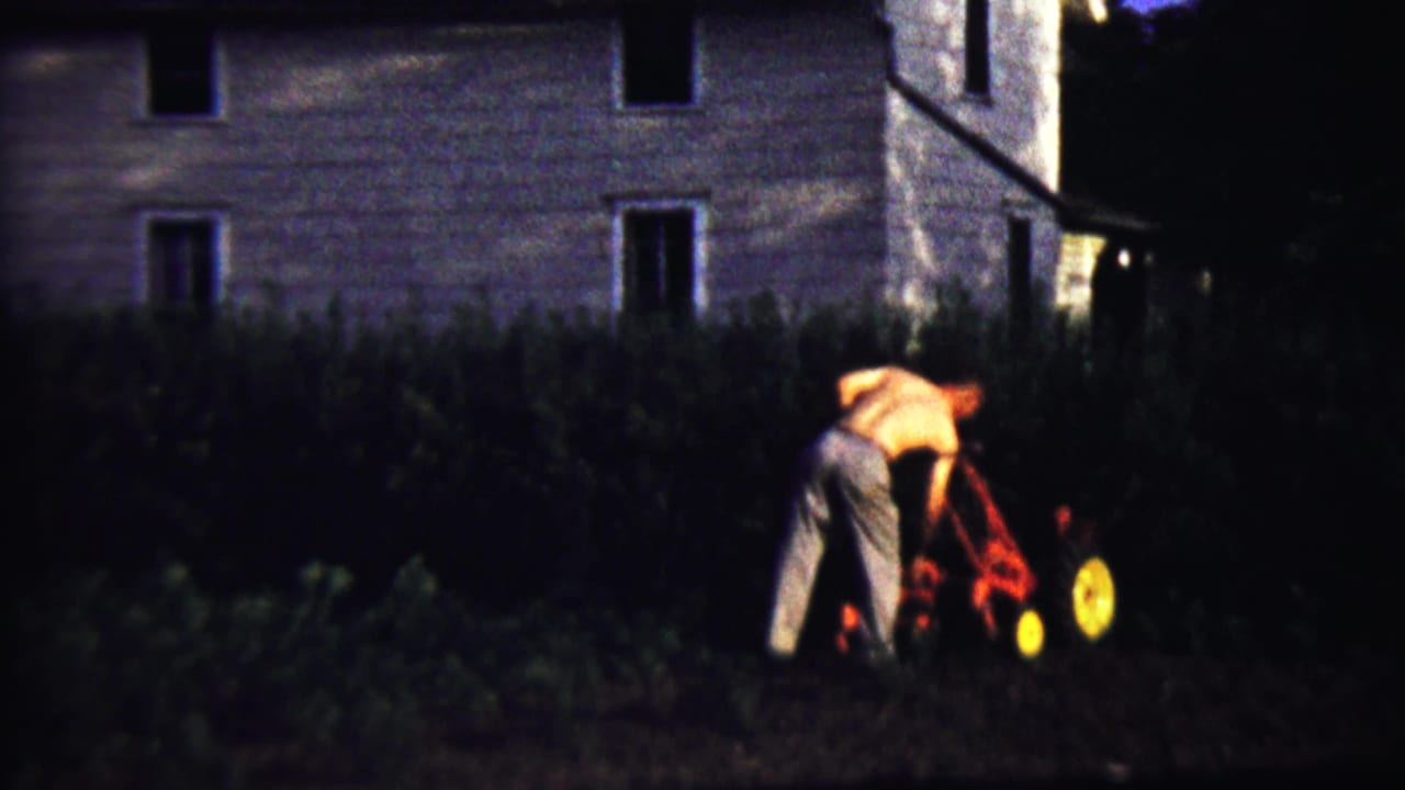 At the end of the clip, the man bends toward the red tiller in the dark garden, with the house exterior and foliage behind him.