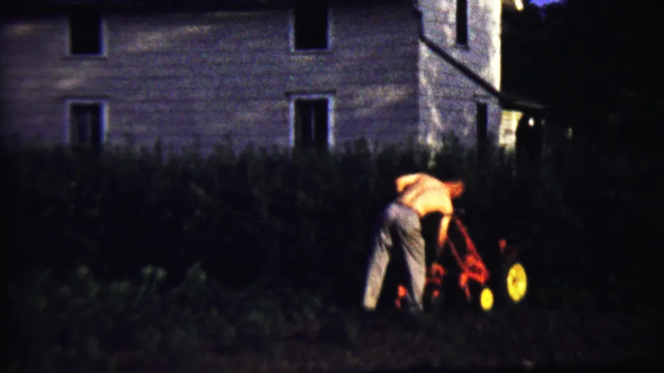 At the end of the clip, the man bends toward the red tiller in the dark garden, with the house exterior and foliage behind him.