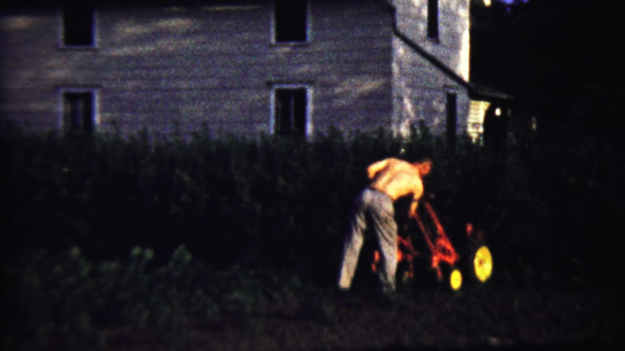 Shirtless man pushing a red lawnmower through tall grass near a house.