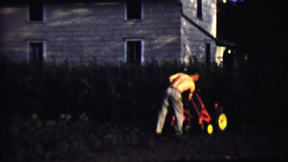 1958 Saint Francis, Kansas home-movie frame showing a shirtless man mowing a grassy yard beside a house.