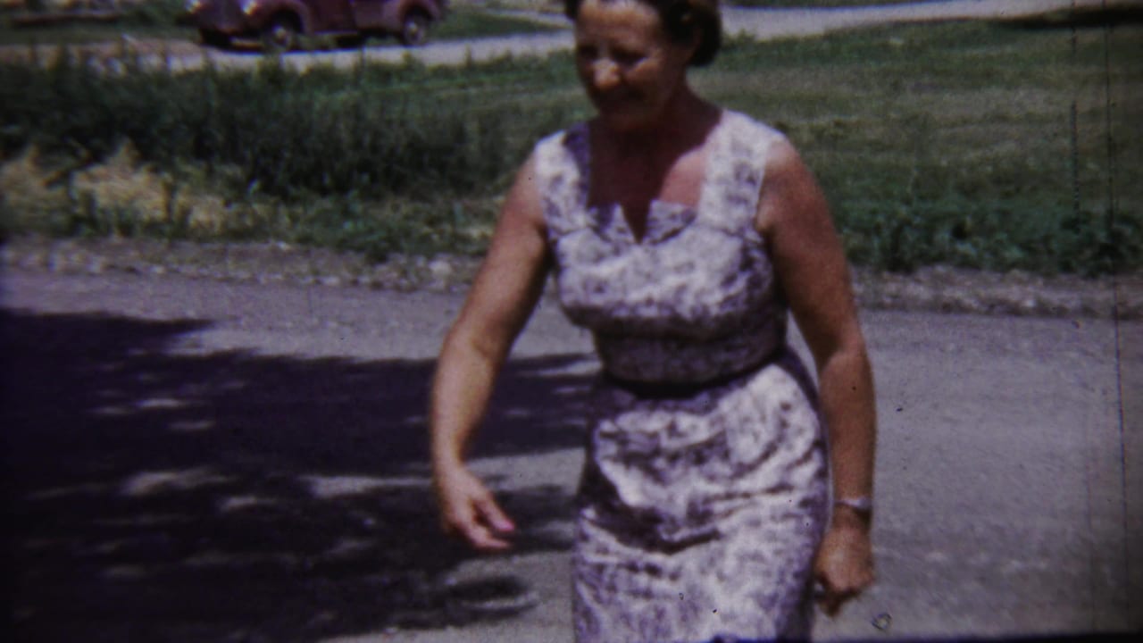 1958 Saint Francis, Kansas frame showing a woman walking across pavement with grass and a parked car in the background.