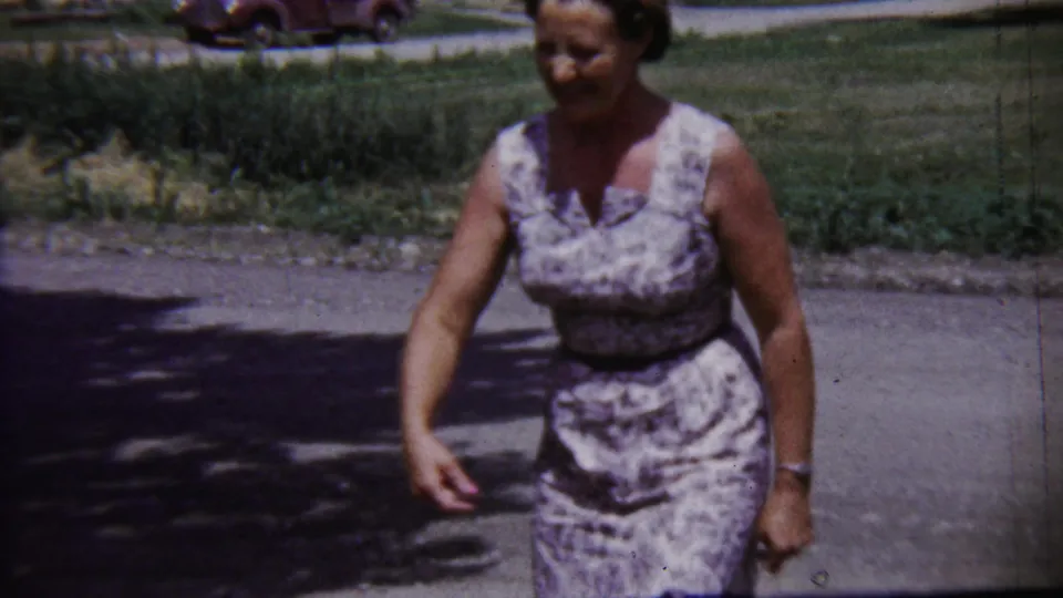 1958 Saint Francis, Kansas frame showing a woman walking across pavement with grass and a parked car in the background.