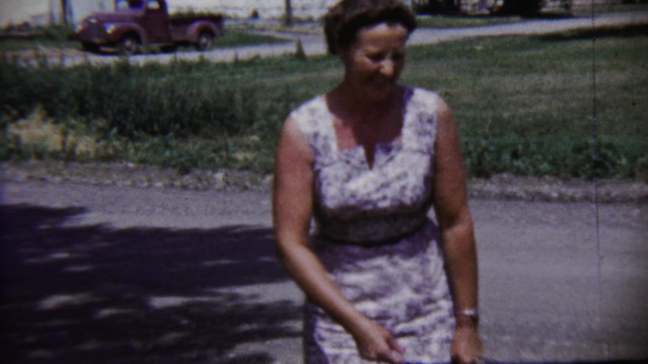 Woman in a patterned sleeveless dress standing on a sunlit street with grass and a red pickup behind her.