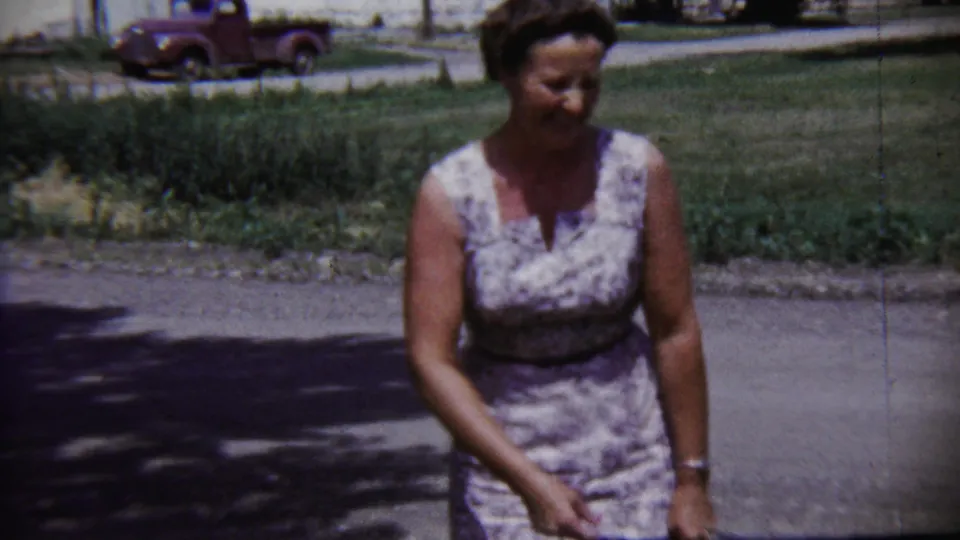 Adult woman in a patterned sleeveless dress on a quiet Saint Francis, Kansas street in 1958, with lawn and a red pickup in the background.