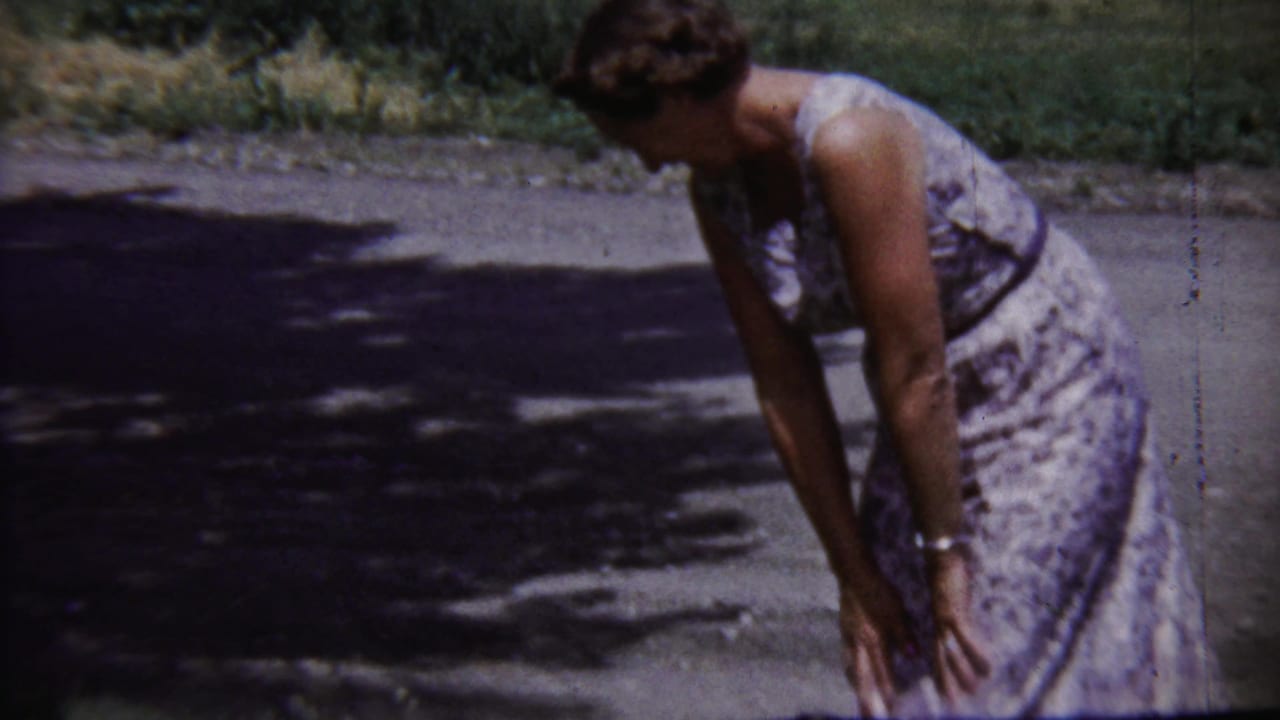 Adult woman in a patterned dress bends forward on a quiet Saint Francis, Kansas street in the 1958 home-movie footage.
