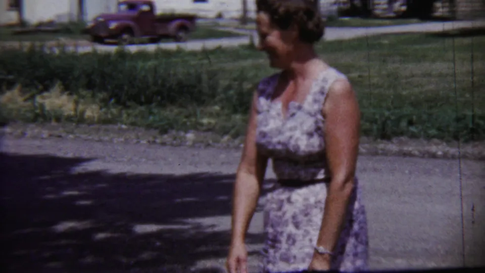 Adult woman turns in profile while walking along a quiet Saint Francis, Kansas street, with a red pickup and lawn behind her.
