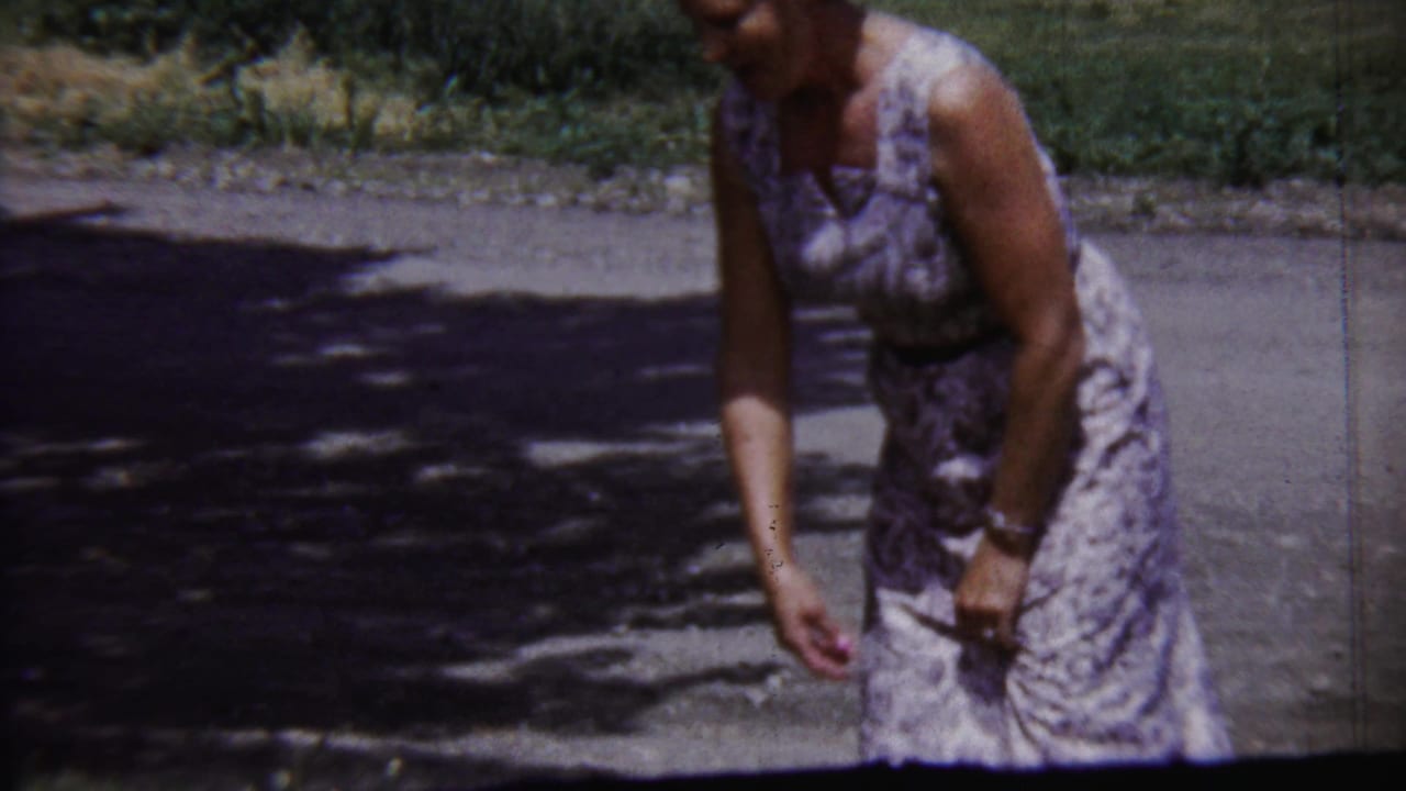 Adult woman bends near the edge of a paved Saint Francis, Kansas street, framed by grass and mottled tree shade.