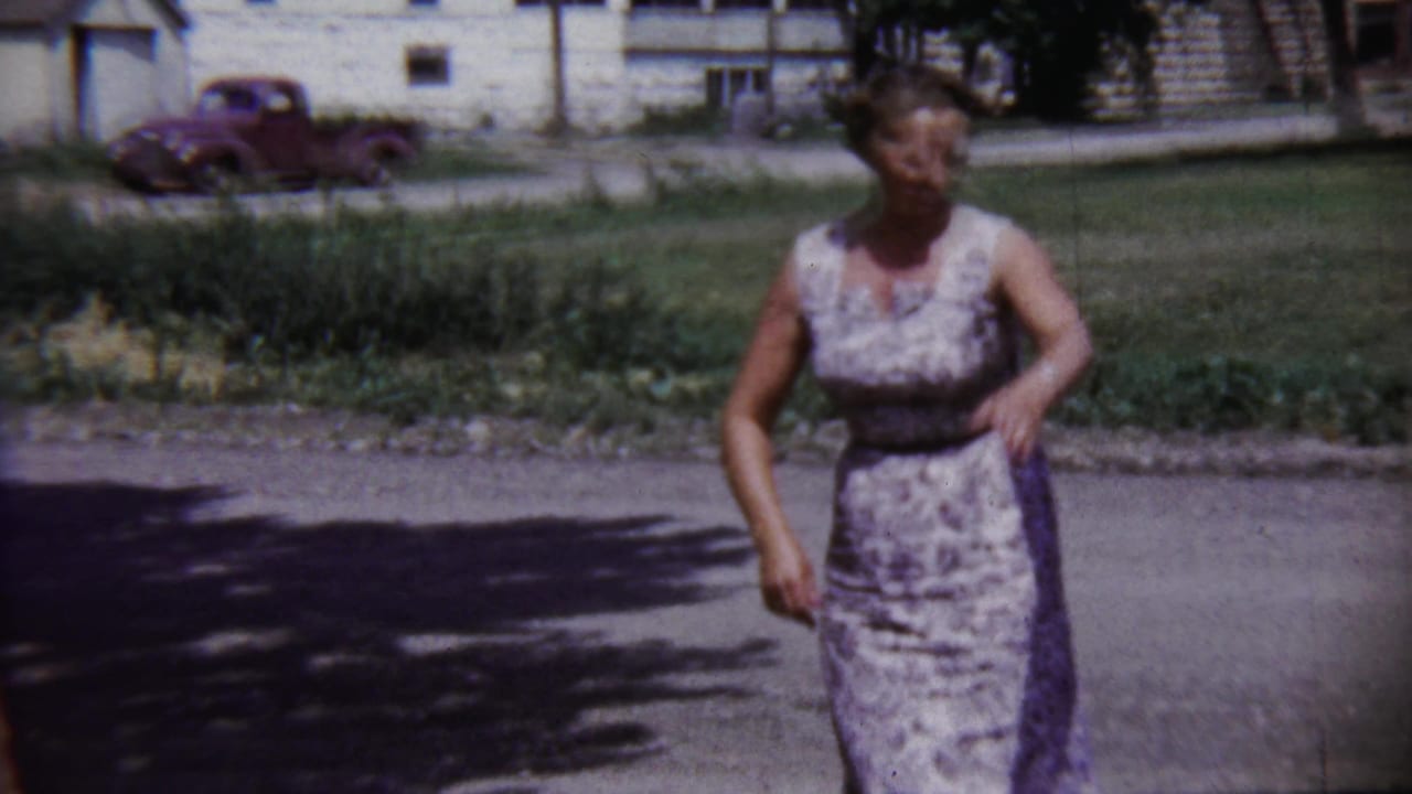 The woman stands on a quiet Saint Francis street, with an older red pickup, lawn, and white house visible in the background.