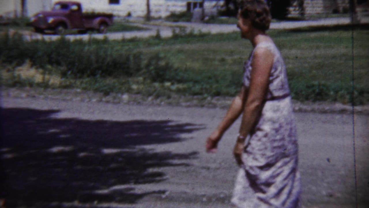 Seen in profile, the woman moves along the street in Saint Francis with a lawn, houses, and older pickup behind her.