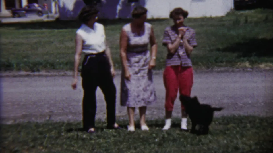 Three young women in a Saint Francis yard face the camera while a small black puppy stands near the woman in red capri pants.