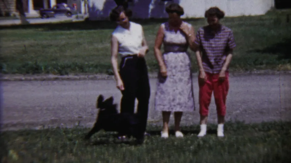 The puppy appears near the feet of three young women in a 1958 Kansas residential yard, with houses and a street behind them.