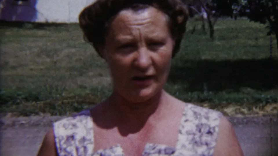 Woman facing a handheld camera outdoors in 1958 Saint Francis, Kansas, with lawn and house details behind her.