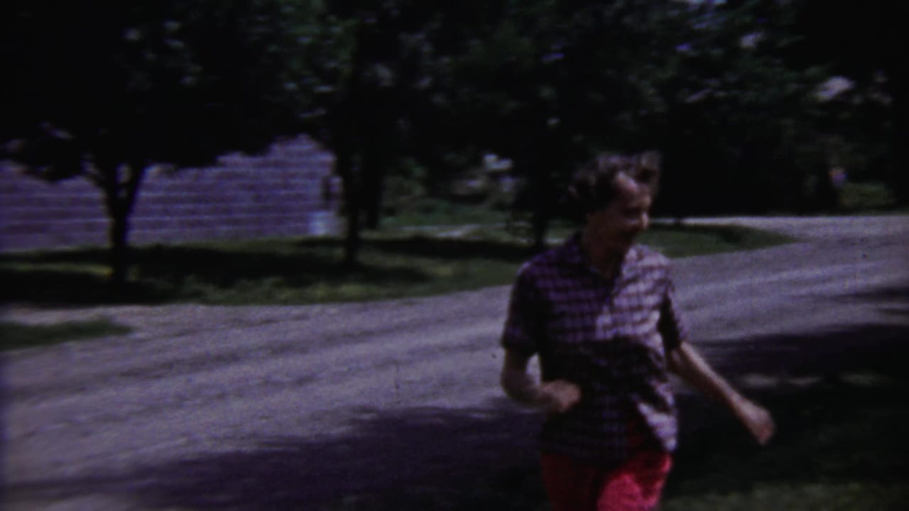 Young person in a plaid shirt and red pants runs across a residential yard in Saint Francis, Kansas, home-movie footage.