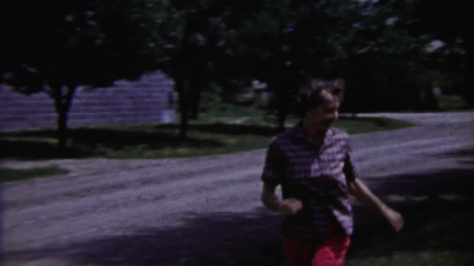 Young person in a plaid shirt and red pants runs across a residential yard in Saint Francis, Kansas, home-movie footage.
