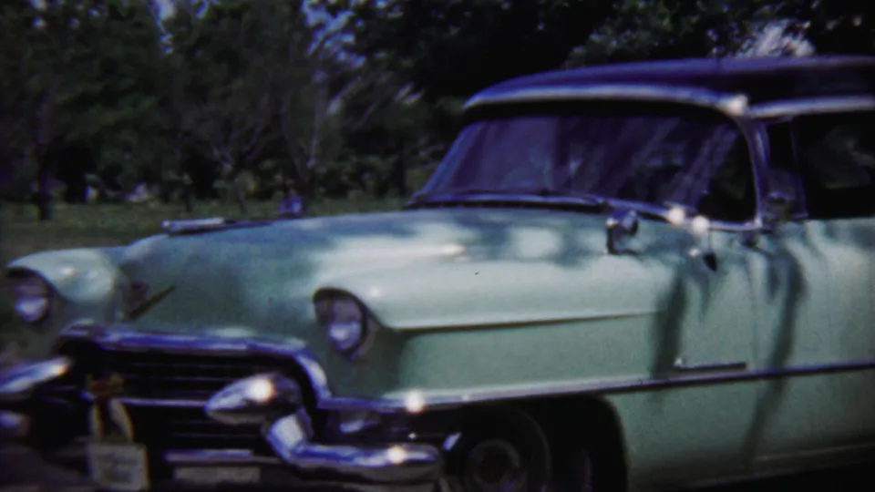 Pale green mid-century sedan in Saint Francis, Kansas, seen in 1958 8mm footage with trees casting shade across the hood and windshield.