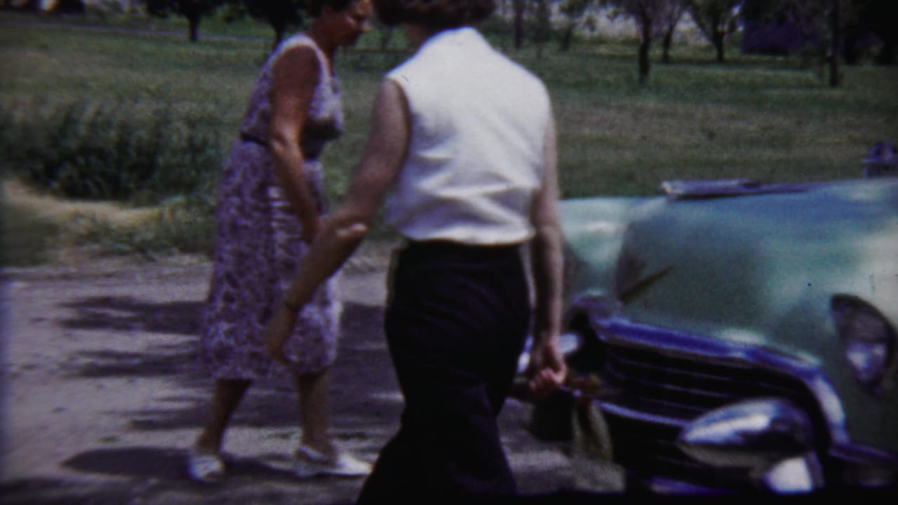 Two adults in period clothing walk past the pale green sedan in the 1958 Saint Francis, Kansas home-movie scene.