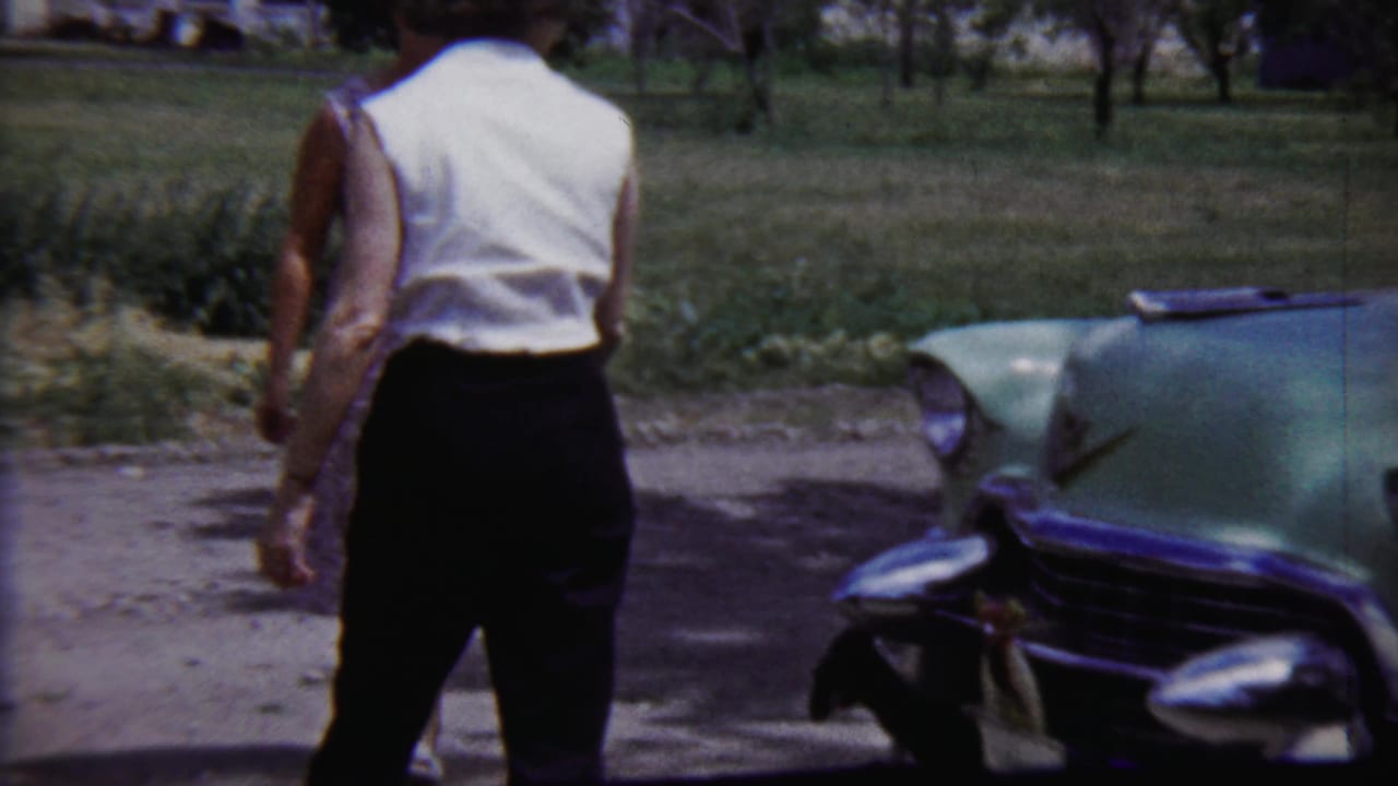 Woman in a white sleeveless blouse stands beside the front of a pale green car.