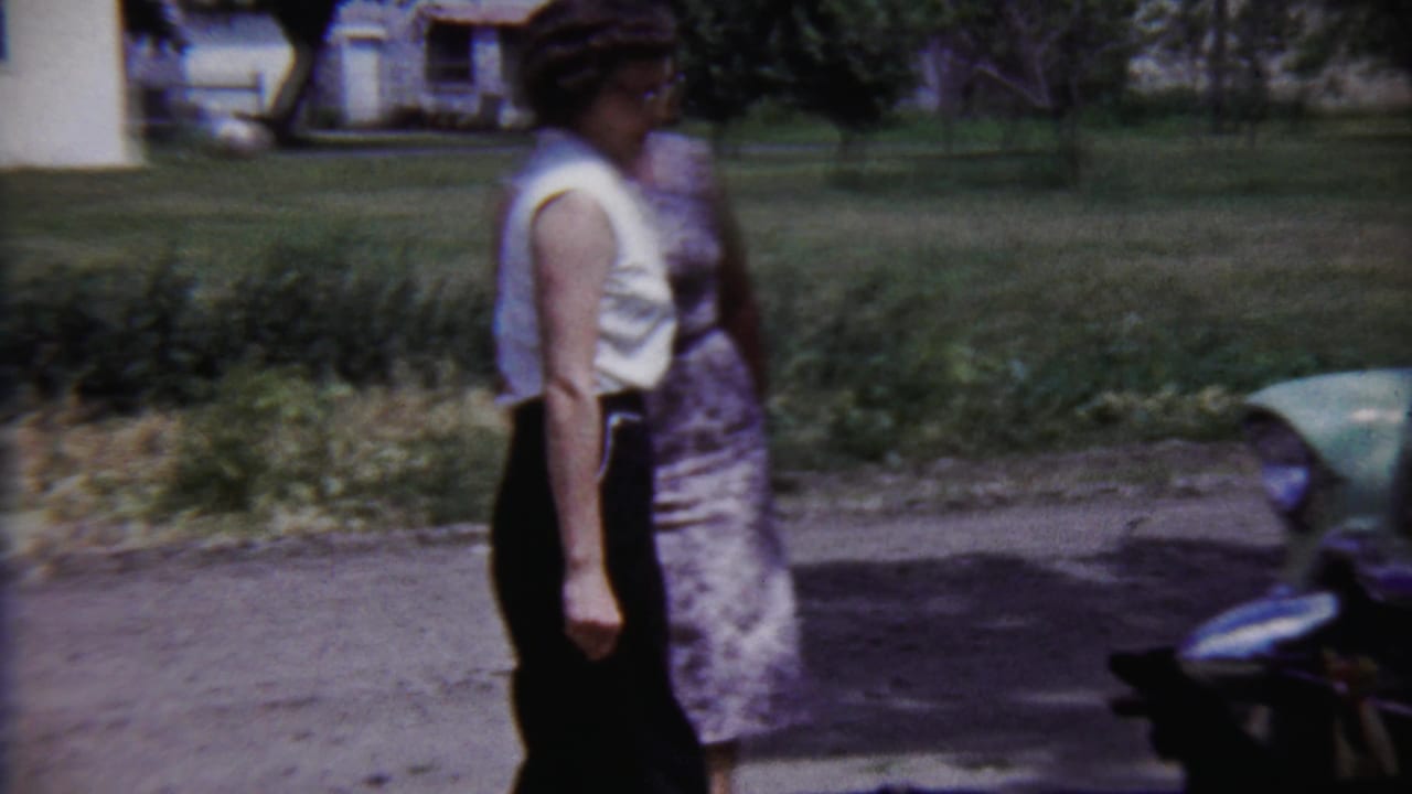 Two women in 1950s attire move past a pale green automobile in an outdoor residential setting in Saint Francis, Kansas.