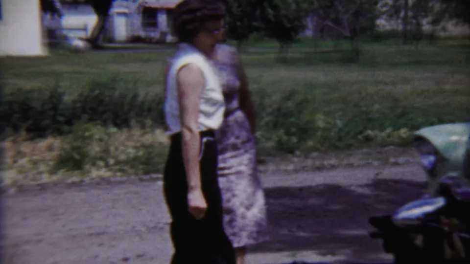 Two women in 1950s attire move past a pale green automobile in an outdoor residential setting in Saint Francis, Kansas.