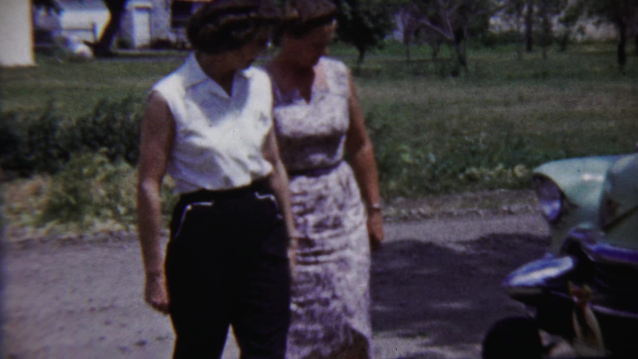 In this 1958 home-movie frame, two women stand by a light green car with lawn and trees behind them.